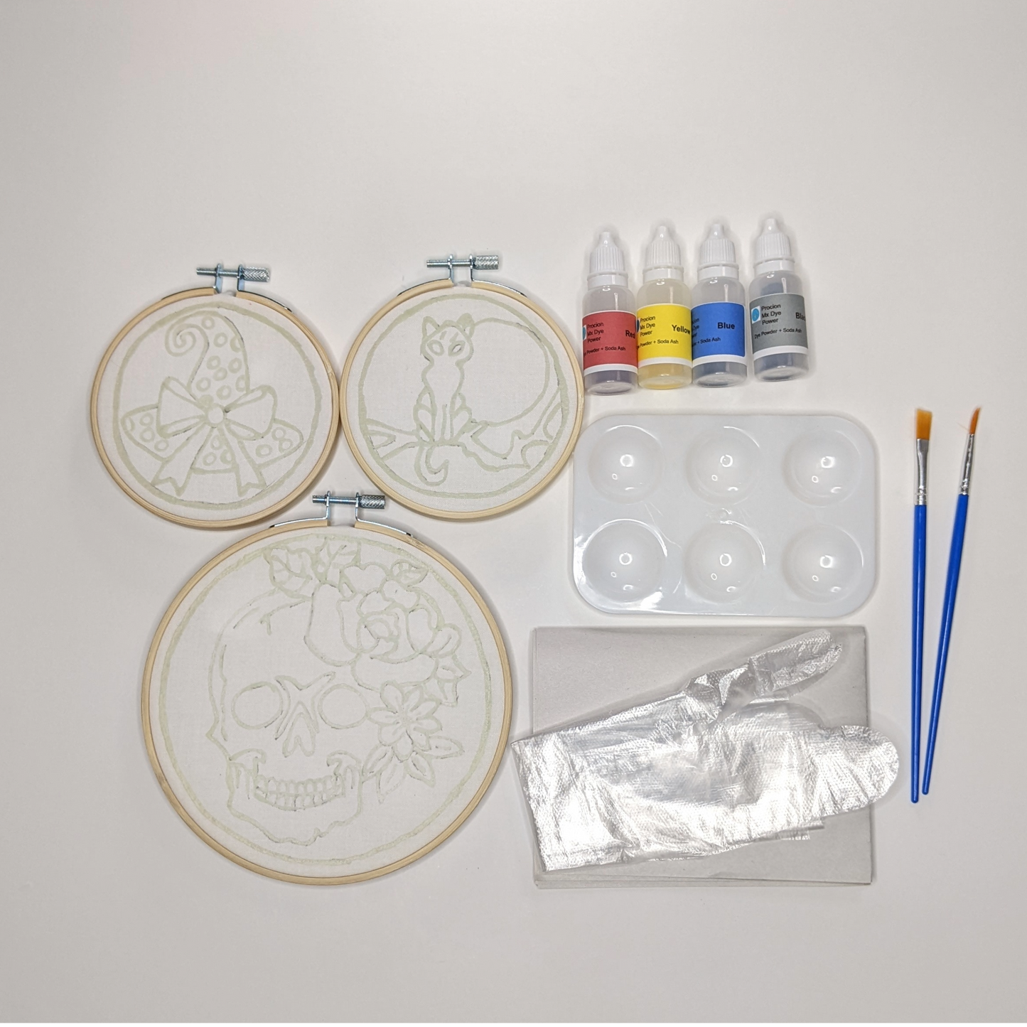 Three hoops with pre-waxed batik designs of a skull with rose and flowers, a moonlit cat on a branch and a witch's hat with a bow  alongside bottles of fabric dyes, palette, brushes, gloves and newsprint.