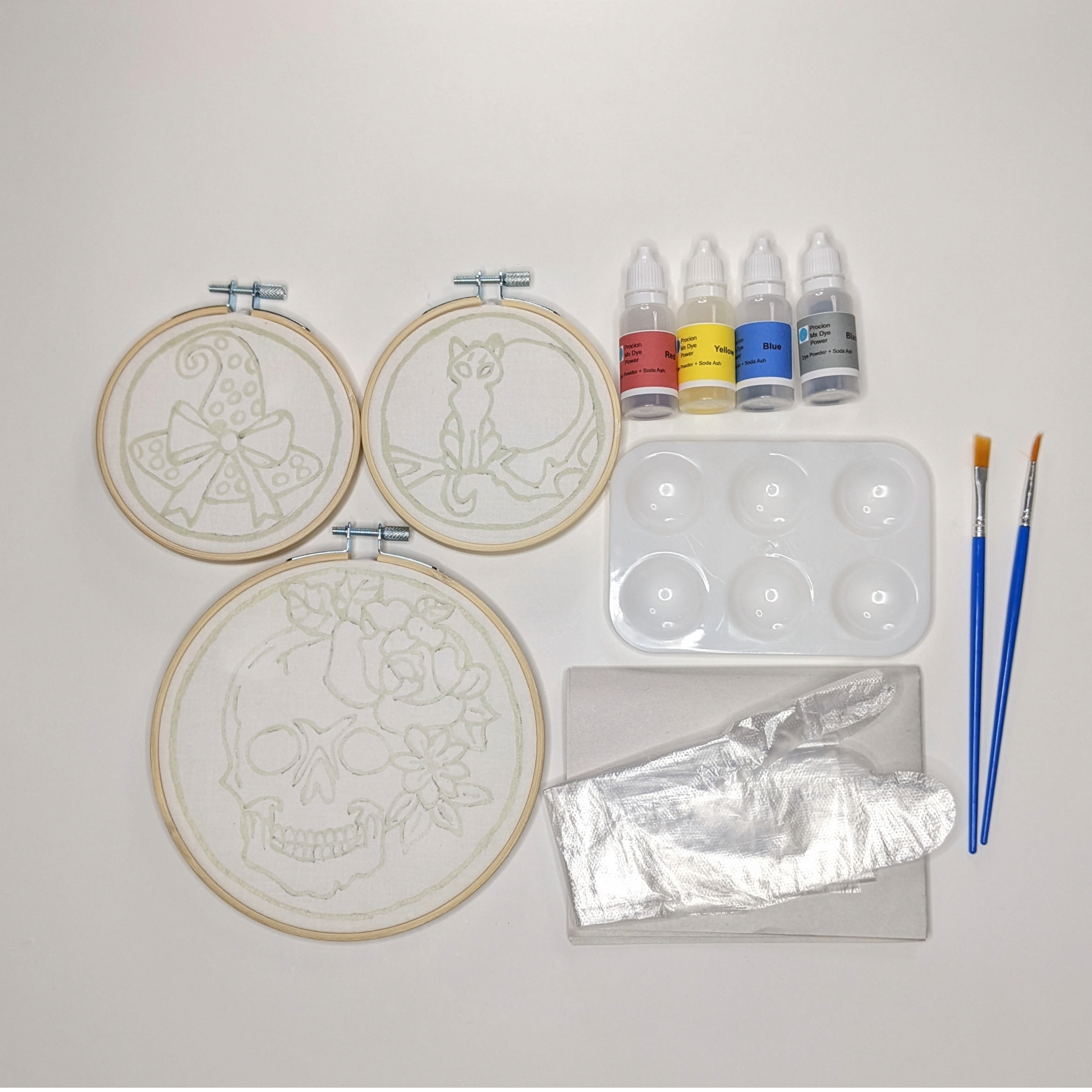 Three hoops with pre-waxed batik designs of a skull with rose and flowers, a moonlit cat on a branch and a witch's hat with a bow  alongside bottles of fabric dyes, palette, brushes, gloves and newsprint.